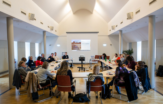Workshop im Black History Month, 19 Frauen und Männer sitzen an U-förmig aufgestellten Tischen, im Hintergrund eine Präsentation auf einer Leinwand